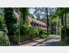 Ein schmaler Weg führt entlang einer Reihe von niedrigen Reihenhäusern inmitten eines grünen, bewaldeten Wohngebiets. Die Häuser haben rote Ziegeldächer mit kleinen Dachgauben und sind von vielen Bäumen, Sträuchern und Blumenbeeten umgeben.