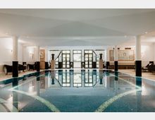 Eleganter Innenpool in einem Spa-Bereich mit blau gefliester Wasserfläche, flankiert von weißen Säulen und Liegestühlen. Im Hintergrund befinden sich große Fenster mit Glastüren, die Tageslicht in den Raum lassen, sowie dekorative Elemente wie Vasen und Bilder.