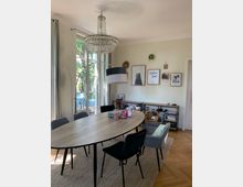 Esszimmer mit einem ovalen Holztisch, umgeben von schwarzen und grauen Stühlen, auf einem Teppich mit Holzfußboden. Eine glitzernde Kristall-Deckenleuchte hängt über dem Tisch, während an der hellgrünen Wand gerahmte Bilder und Dekorationen angebracht sind. Im Hintergrund stehen Regale und eine Fensterfront mit Blick ins Freie.