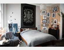 Schlafzimmer mit einem Holzbett und grauer gesteppter Bettdecke, an der Wand ein großes schwarzes Wandtuch mit Wappenmotiv. Rechts eine Wand mit sichtbarer Patina und einer Sammlung von gerahmten Fotos, links ein Sessel mit grauer Decke. Eine moderne Stehlampe und eine Kugelleuchte ergänzen die Einrichtung.