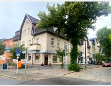 Straßenszene mit einem beigefarbenen Gebäude mit Satteldach, in dem sich eine Apotheke im Erdgeschoss befindet. Umgeben von Bäumen, Kopfsteinpflaster und Straßenschildern, darunter ein Taxi-Haltestellenschild, sowie geparkte Autos am Straßenrand.