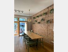 Ein lichtdurchfluteter Raum mit einer langen Holztischgruppe und grünen Stühlen auf einem Holzboden. An der freigelegten Ziegelwand hängen zahlreiche Topfpflanzen an einem Gitter, während eine Glastür mit Holzrahmen den Blick nach draußen freigibt.