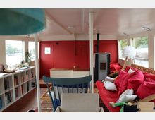 Wohnbereich eines Hausboots mit weißen Wänden und roten Akzenten. Zu sehen sind ein rotes Sofa mit Kissen, ein Holzofen, ein Regal mit Büchern sowie Fenster, die den Blick aufs Wasser ermöglichen.