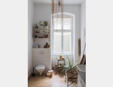 Helles Badezimmer mit Holzboden, einem kleinen Wand-WC links und einem großen Fenster mit halbtransparentem Vorhang. Dekorative Elemente umfassen Pflanzen, hängende Zweige von der Decke und Regalböden mit Keramik sowie Körbe und ein Rattanstuhl.