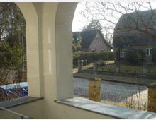 Blick von einer überdachten Terrasse mit weißen Rundbögen auf eine gepflasterte Straße und gegenüberliegende Einfamilienhäuser mit Schrägdächern. Im Vordergrund sind Teile eines Geländers und ein blattloser Baum sichtbar.