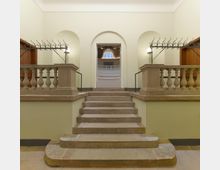 Treppenaufgang in einem historischen Gebäude mit steinernen Stufen, flankiert von Balustraden und Kleiderhakenreihen. Im Hintergrund ein Türbogen, der den Blick auf einen Saal mit großer Fensteröffnung und dekorativen Elementen freigibt.