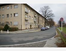 Ecke einer Wohnstraße mit einem dreistöckigen Wohngebäude aus beigefarbenem Putz und mehreren Balkonen. Links ein kleiner Grünstreifen mit Büschen, rechts die Straße mit geparkten Autos, einem Rasenstreifen und einem Verkehrsschild mit Halteverbot.