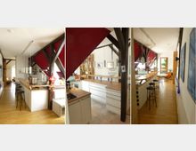 Weitläufiger Küchen- und Wohnbereich in einem Dachgeschoss mit sichtbaren Holzträgersystemen, weißen Wänden und roten Akzentelementen. Die Küche verfügt über Holzarbeitspplatten, eine Theke mit Barhockern und moderne Hängelampen, während im Hintergrund Sitzgelegenheiten und eine Tür zum Außenbereich erkennbar sind.