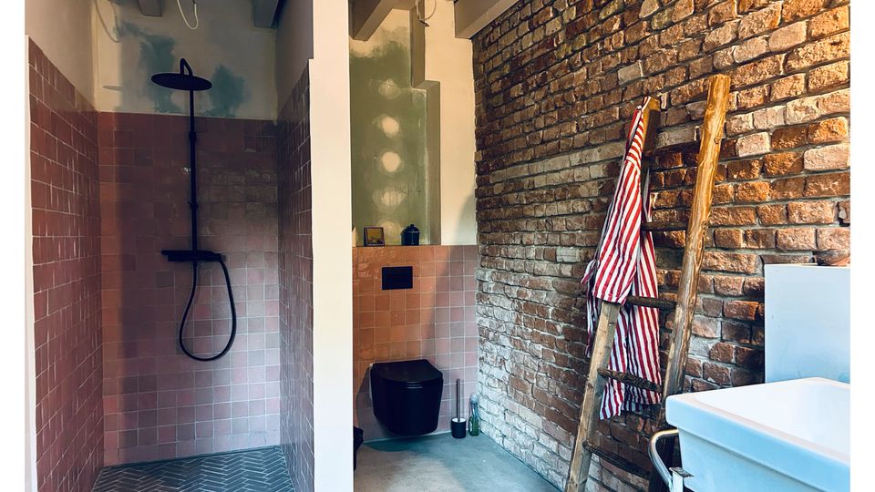 Innenansicht eines schmalen Badezimmers mit freiliegender Ziegelsteinwand, terrakottafarbenen Bodenfliesen und einer Dusche mit schwarzer Armatur. Eine Holzleiter mit gestreiftem Handtuch lehnt an der Wand; daneben stehen ein schwarzes Waschbecken und eine Badewanne.