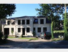 Eine moderne zweistöckige Villa mit hellbeiger Fassade, schwarzen Fenstern und einem dunkelblauen Walmdach, umgeben von Bäumen und einem gepflegten Garten. Auf der Auffahrt vor dem Haus steht ein geparktes Auto.