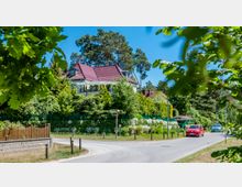 Wohnhaus mit rotem Ziegeldach und großer Terrasse, umgeben von dichten Bäumen und Sträuchern. Im Vordergrund eine schmale asphaltierte Straße mit fahrenden Autos und ein Holzzaun, umgeben von grünem Gelände.