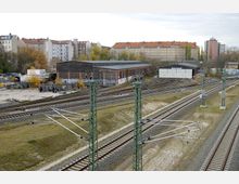 Bahngelände mit mehreren Gleisen und Oberleitungsmasten im Vordergrund, im Hintergrund stehen flache Industriebauten sowie Wohnhäuser mit roten Ziegeldächern und hellen Fassaden. Die Umgebung zeigt eine Mischung aus urbanen Strukturen und herbstlicher Vegetation.