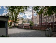 Hinterhof mit einem kleinen, umzäunten Bolzplatz, umgeben von Bäumen, Backsteinmauern mit Graffiti und angrenzenden mehrstöckigen Wohn- und Geschäftsgebäuden; im Hintergrund ein hoher Schornstein.