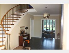 Eingangsbereich eines Hauses mit einer geschwungenen Holztreppe links, einem kleinen Tisch mit Lampe und Dekoration an der rechten Wand und Blick in ein angrenzendes Esszimmer mit großem Fenster und Tisch mit Stühlen. Eine kugelförmige Deckenleuchte hängt in der Mitte des Raumes.