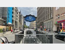 Straßenansicht von Berlin mit dem U-Bahneingang "Kochstraße – Checkpoint Charlie" im Vordergrund, umgeben von mehrstöckigen Gebäuden, darunter eine mit Baugerüsten bedeckte Fassade. Fahrräder sind an einem Ständer geparkt, und Passanten sowie Autos beleben die Szene.