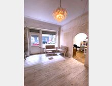 Ein heller Raum mit Holzdielenboden, Vintage-Sitzmöbeln und einer dekorativen Deckenleuchte. Eine große Fensterfront mit Tür führt zur Straße, während ein offener Rundbogen den Blick auf einen angrenzenden Raum freigibt.