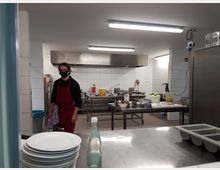 Große, professionelle Küche mit weiß gefliesten Wänden und Edelstahl-Arbeitsflächen. Im Vordergrund sind gestapelte Teller und eine Glasflasche zu sehen, während im Hintergrund eine Person mit roter Schürze und schwarzer Gesichtsmaske neben Küchengeräten und Utensilien steht.