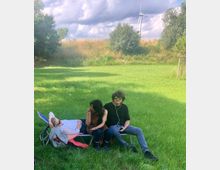 Eine Wiese mit drei Personen, die auf einem Liegestuhl sitzen oder liegen; im Hintergrund sind Bäume, eine kleine Anhöhe und ein Windrad unter bewölktem Himmel zu sehen.