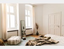 Helles Schlafzimmer mit großen Fenstern, einem weißen Holztürrahmen und minimaler Dekoration. Im Raum sind ein flauschiger Hocker, ein großer Spiegel, eine grüne Vase mit Trockenblumen und eine Pelzdecke auf dem Bett zu sehen.