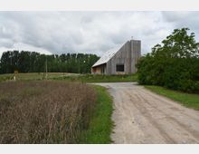 Modernes Gebäude aus Holz mit asymmetrischem Satteldach, umgeben von einer ländlichen Landschaft mit Feldern, Bäumen und einem unbefestigten Weg im Vordergrund. Der Himmel ist bewölkt, und Vegetation dominiert die Umgebung.