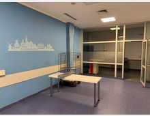 Ein einfacher Raum mit blauem Boden und blauer Wand, auf der eine stilisierte Skyline von Flensburg abgebildet ist. Im Raum befinden sich ein weißer Tisch, ein Metallrollwagen und eine Glaswand mit einer Tür, die in einen angrenzenden Bereich führt.