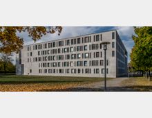 Modernes vierstöckiges Gebäude aus Beton mit vielen rechteckigen Fenstern, umgeben von einer Rasenfläche und Herbstbäumen. Ein gepflasterter Weg mit Laternen führt am Gebäude entlang.