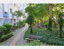 Grüne Innenhofanlage mit gepflasterten Wegen, Sitzbank, Fahrrädern und viel Efeu bewachsenen Flächen. Im Hintergrund ein mehrstöckiges Wohnhaus mit weißen und blauen Fassadenelementen.