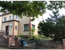 Zweistöckiges Wohnhaus mit hellgelber Fassade und roten Backsteinelementen, einem kleinen Vorgarten mit Sträuchern und Büschen sowie einem Metallzaun und Treppenaufgang zur Eingangstür. Im Vordergrund ragen Zweige eines Baumes ins Bild.