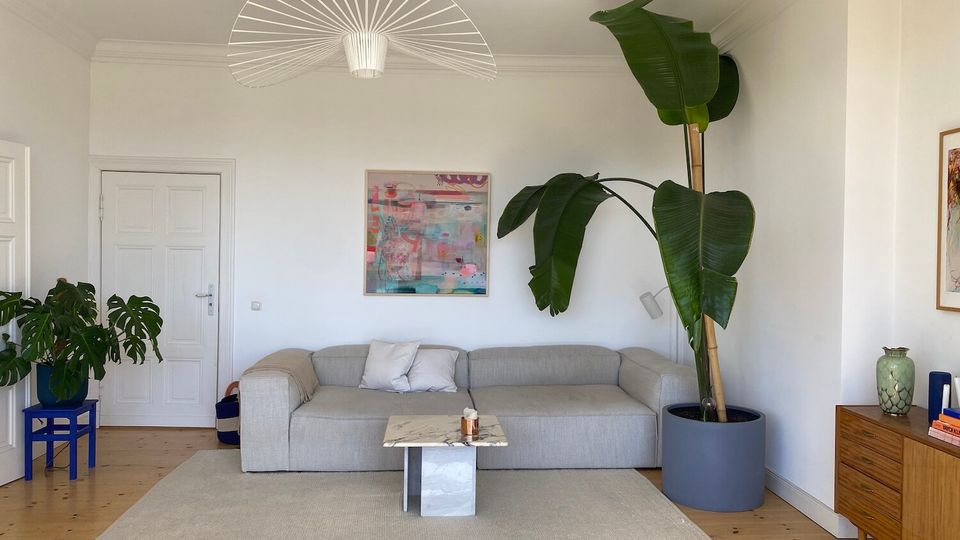 Bright, minimalist living room with white walls, wood floors, and a large gray sectional sofa on a light area rug. A small marble-topped coffee table sits in front of the sofa, with a large potted plant, wall art, and a sculptural ceiling light above.