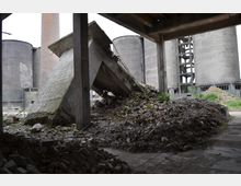 Ruine eines Industriegebäudes mit Betontrümmern, Schutthaufen und überwuchertem Gras im Vordergrund. Im Hintergrund sind runde, graue Silos und ein hoher Schornstein zu sehen.