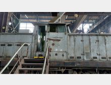 Alte, rostige Lokomotive in einer Werkhalle mit freiliegenden Stahlträgern und einer großen Fensterfront im Hintergrund. Eine Metalltreppe führt zur Fahrerkabine, deren Tür offensteht.