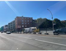 Straßenszene mit einem mehrstöckigen roten Wohngebäude auf der linken Seite, einer U-Bahn-Zugangsstation mit einem Schild "U" und vorbeifahrenden Fahrzeugen, darunter ein gelber Linienbus. Im Hintergrund sind Bäume und ein blauer Himmel zu sehen.