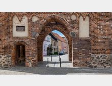 Backsteinbogen eines historischen Stadttors mit Sicht auf eine gepflasterte Straße und bunte Häuserfassaden im Hintergrund. Der Torbogen ist mit gotischen Verzierungen und kleinen, zugemauerten Öffnungen gestaltet.