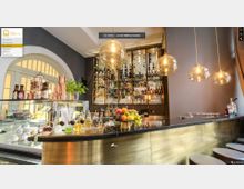 Barbereich mit einem goldfarbenen Tresen und schwarzen Arbeitsflächen, dekoriert mit frischem Obst sowie Flaschen und Gläsern. Im Hintergrund befindet sich ein Regal mit einer großen Auswahl an Getränken, beleuchtet von mehreren goldenen Hängelampen.