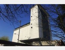 Eine moderne Kirchenfassade mit einem hohen, rechteckigen Kirchturm aus Beton, der eine große Fensterfront aufweist. Die Umgebung zeigt vereinzelt Bäume und blauen Himmel im Hintergrund.