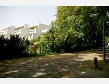 Mehrstöckiges weißes Wohngebäude mit terrassenförmigen Balkonen, umgeben von dichtem Grün und kleinen Bäumen. Im Vordergrund befindet sich eine asphaltierte Straße mit Schatten von Bäumen und einer Straßenlaterne.