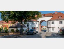 Ein gepflasterter Weg führt durch einen großen weißen Torbogen, der an beide Seiten von Gebäuden mit roten Dächern angebaut ist. Im Vordergrund steht ein parkendes Auto unter einem großen Baum, während im Hintergrund weitere Wohnhäuser mit gelben Fassaden zu sehen sind.