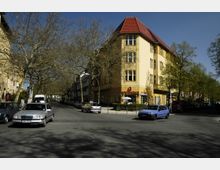 Straßenecke mit einem gelben Gebäude im Hintergrund, das ein rotes Dach und mehrere Fenster hat; im Erdgeschoss befindet sich eine Apotheke mit rotem Schriftzug. Die Straße ist von kahlen Bäumen gesäumt, und mehrere parkende sowie fahrende Autos sind sichtbar.
