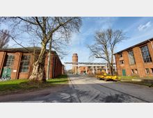 Industriegelände mit Backsteingebäuden, großen Fenstern und einem Wasserturm im Hintergrund. Über die Straße verläuft eine weiße Metallbrücke, und am Rand stehen Bäume sowie ein gelber Transportwagen.