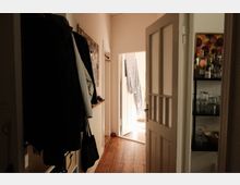 Schmaler Wohnungsflur mit Holzdielen, heller Wand und Garderobe an der linken Seite. Rechts steht eine geöffnete Tür mit Glaseinsätzen neben einem Regal mit Flaschen und Gläsern; am Ende führt eine offene Tür in einen hell erleuchteten Raum.