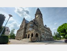 Historisches Gebäude aus Sandstein mit einem hohen Turm, Arkaden am Eingangsbereich und verzierter Fassade. Im Vordergrund führen Treppenstufen zum Haupteingang, während im Hintergrund Bäume und ein bewölkter Himmel zu sehen sind.