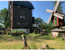Holzmühle auf einem grünen Gelände mit Gartenbeeten und kleinen Holzhütten; daneben eine bunte, dekorative Windmühle mit rot-grün gestreiften Flügeln. Im Hintergrund stehen Bäume und weitere Gebäude.