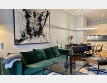 Modernes Wohnzimmer mit einer grünen Samtcouch, einem Couchtisch aus Glas und einem schwarzen Ledersessel. Im Hintergrund eine offene Küche mit weißen Schränken und dunkler Arbeitsplatte, ein abstraktes Kunstgemälde an der Wand ergänzt die minimalistische Gestaltung.