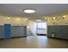 Treppenhaus eines U-Bahn- oder Bahnhofgebäudes mit hellblauen Fliesenwänden, einer blauen Tür auf der linken Seite, mehreren Runddeckenleuchten und Graffiti an den Wänden. Ein großes Fenster im Hintergrund lässt Tageslicht in den Raum fallen.