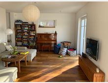 Wohnzimmer mit Holzboden, grauem Ecksofa und rustikalem Couchtisch. Im Hintergrund stehen ein Regal mit Gesellschaftsspielen, ein antikes Klavier und ein Tischkicker; daneben eine Glastür, die auf eine Terrasse führt. Ein Flachbildfernseher hängt an der rechten Wand.