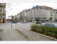 Straßenszene in einer Stadt mit mehrstöckigen Wohn- und Geschäftsgebäuden im Hintergrund, einer Kreuzung mit mehreren Autos, Gehwegen und einem U-Bahn-Zugangsschild mit der Aufschrift „U Wedding“. Im Vordergrund sind ein Fußgängerweg und eine niedrige Metallabsperrung zu sehen.