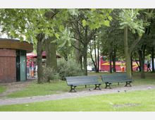 Ein Park mit grüner Wiese, mehreren Bäumen und zwei leeren Holzparkbänken im Vordergrund. Im Hintergrund sind ein gemauertes Gebäude mit roten Sonnenschirmen und Verkaufsständen sowie ein roter Doppeldeckerbus zu sehen.