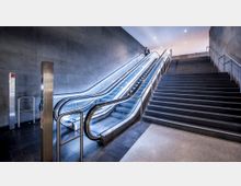 Moderner Innenraum mit zwei beleuchteten Rolltreppen und einer angrenzenden Treppe aus dunklem Stein. Die Wände und der Boden bestehen ebenfalls aus glattem, dunklem Material, der Bereich ist gut beleuchtet.