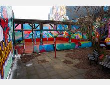 Innenhof mit gepflastertem Boden und Sitzbänken, umgeben von Wänden mit großflächigen, bunten Graffiti-Motiven. In der Mitte steht eine offene, überdachte Plattform mit Holz- und Glasrahmen sowie niedriger, farbig gestrichener Brüstung; daneben ein kahler Baum.