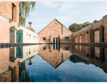Innenhof eines historischen Backsteingebäudes mit symmetrischen, schrägen Dächern und Wänden, die in einem Wasserbecken auf dem Boden spiegeln. Umliegende Mauern aus Ziegeln und Holztüren, flankiert von Bäumen im Hintergrund.